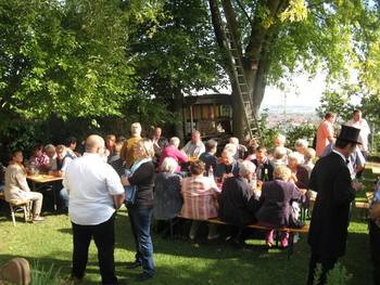 Ökumenischer Abschluss der Kirchweih-Gottesdienste Menschen stehen und sitzen nach den Kirchweihgottesdiensten in einem sommerlichen Garten beisammen, essen und trinken und unterhalten sich
